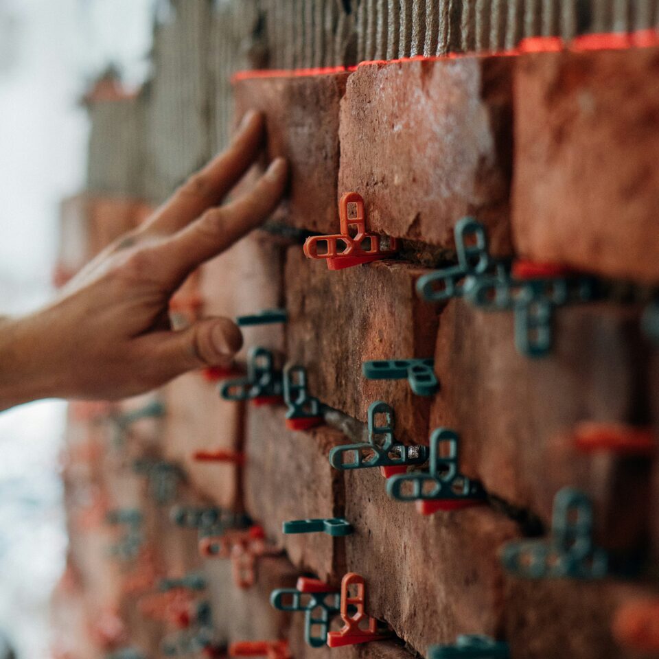 A hand resting against a partly built red brick wall, with coloured plastic spacers set into the unfinished mortar joints.