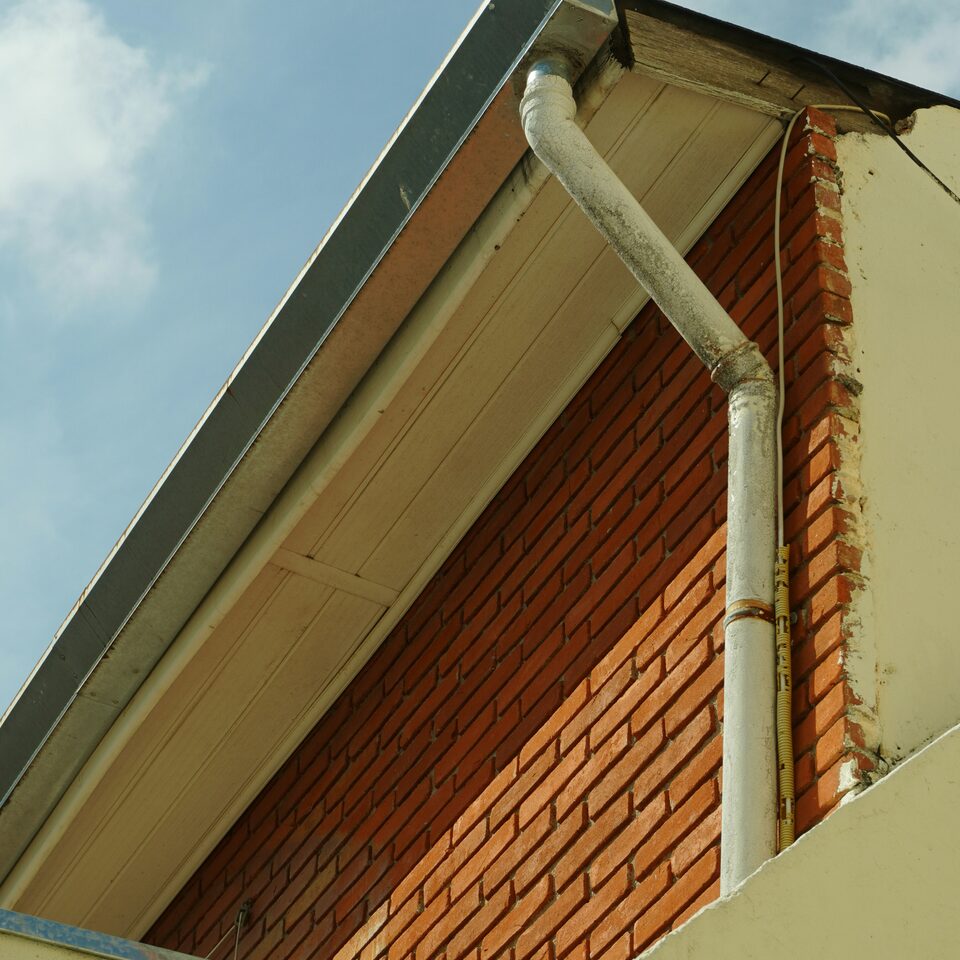 The eaves of a brick building seen from below, showing a painted soffit board, the gutter along its edge and a round downpipe running down the corner.