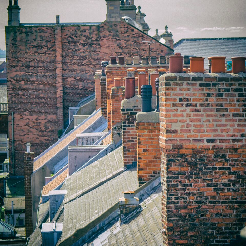 A view across the slate roofs of a terrace, with tall red brick chimney stacks and clay pots standing above the slopes.