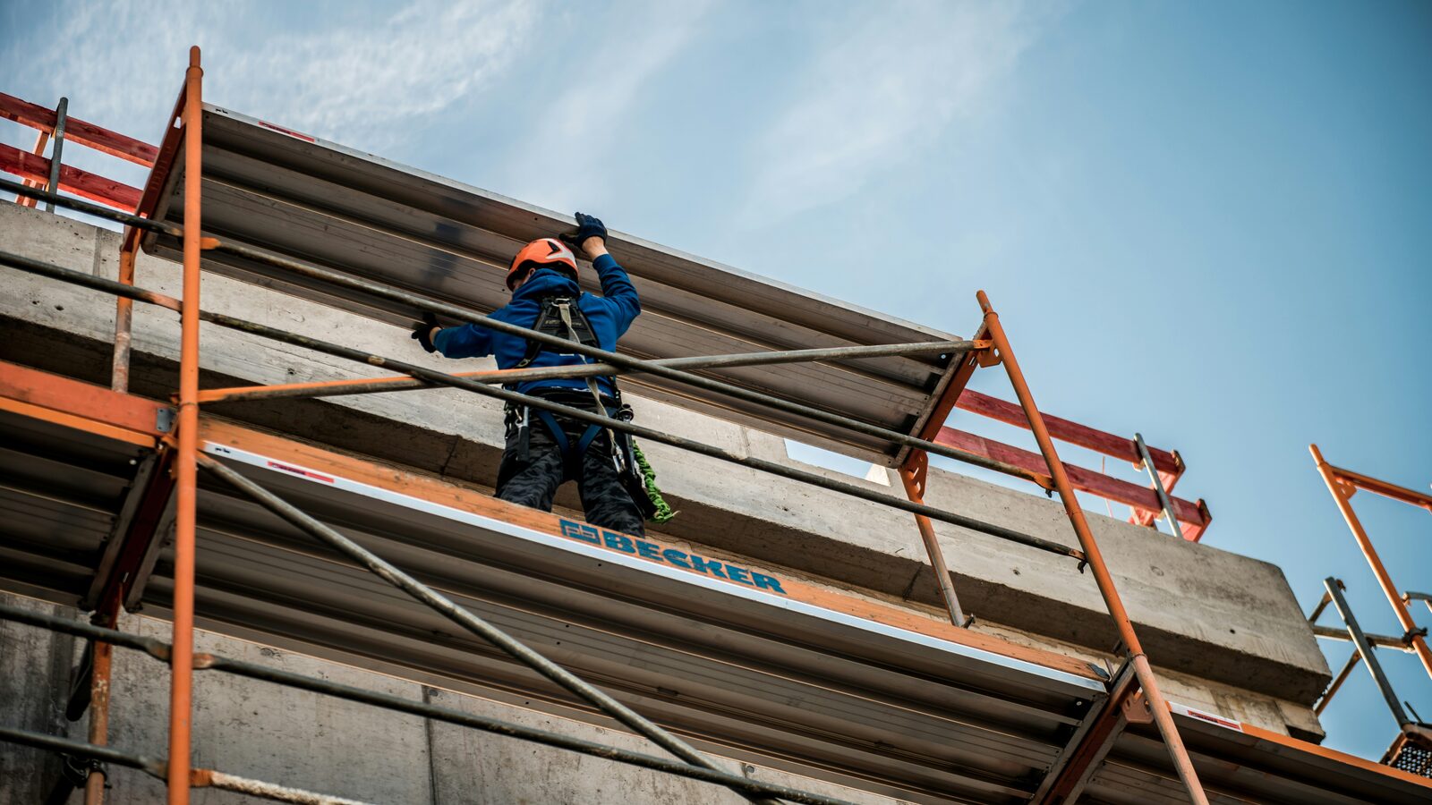 A worker in blue overalls standing on a scaffold platform beside a concrete building under construction, seen from below against a blue sky.
