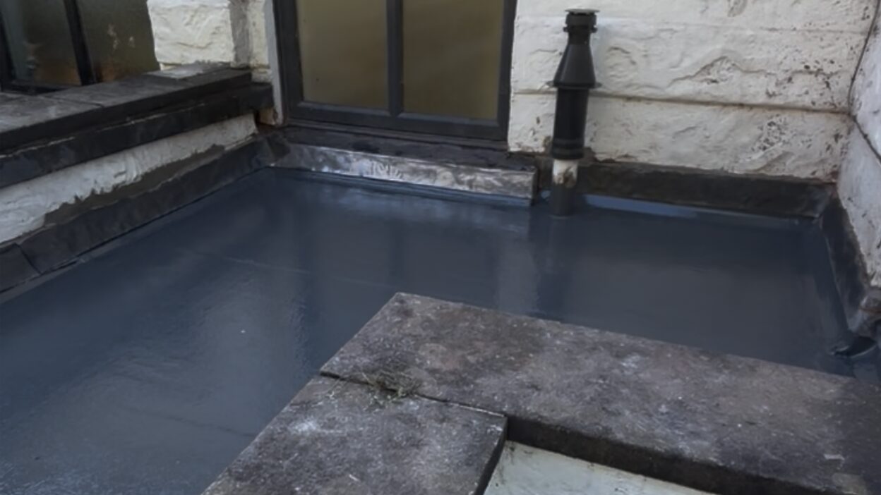 A newly finished grey flat roof between rendered walls, with the waterproof finish turned up at the edges, a metal flashing beneath a window and a vent pipe coming through the roof.