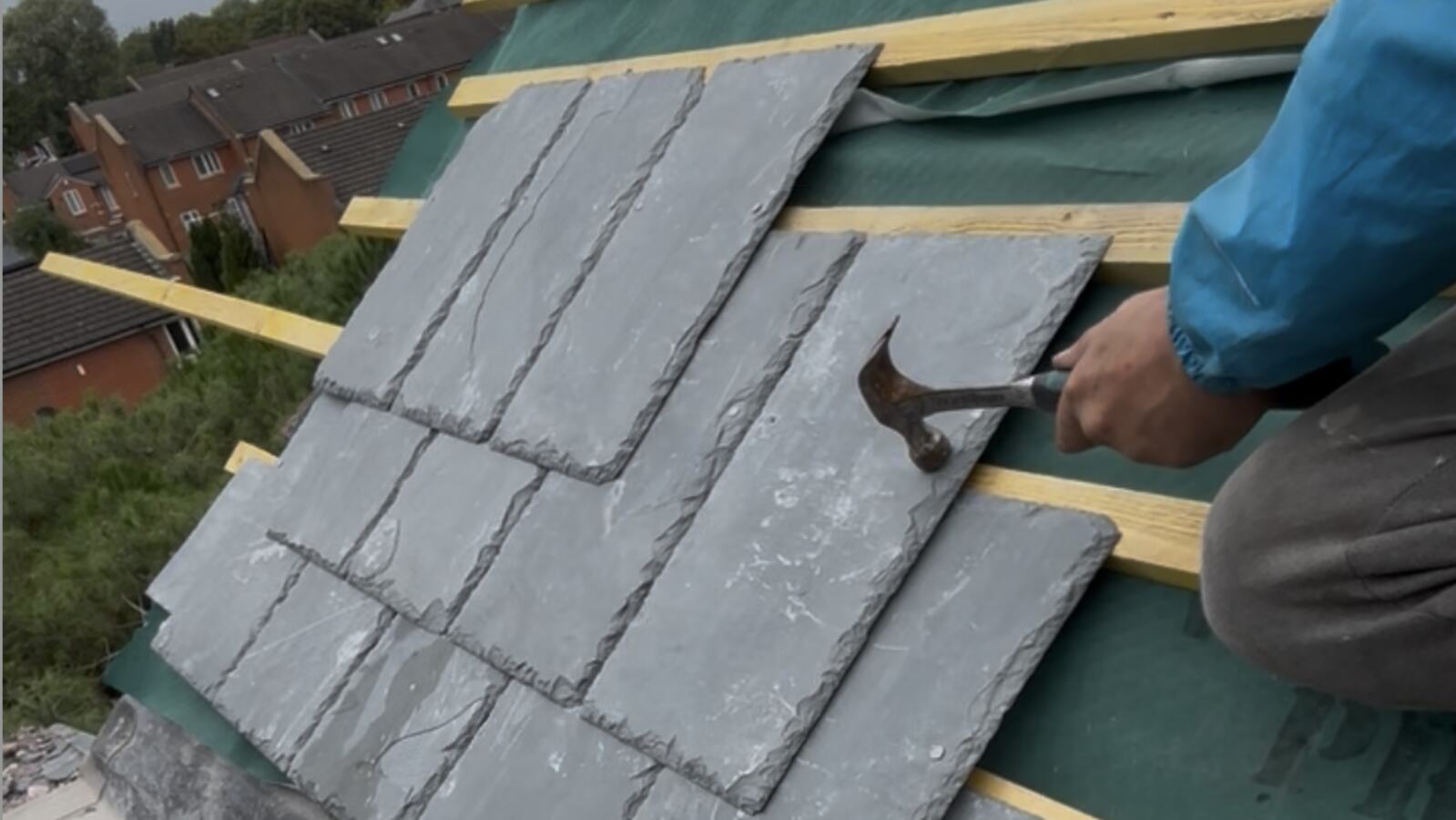 A roofer nailing new natural slates onto fresh timber battens laid over green breathable membrane, with the first courses of slate in place and houses in the background.