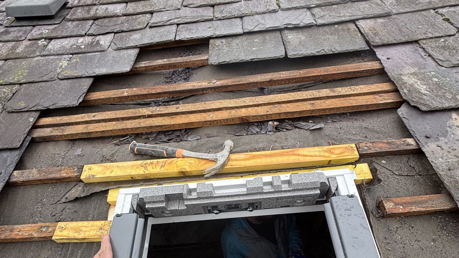 A natural slate roof opened up during work, with slates stripped back to the battens, new timber battens fixed in place, a claw hammer resting on them and the top of a new roof window frame below.