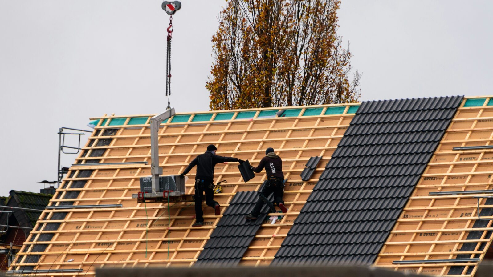 Two roofers working on a steeply pitched roof, laying dark tiles over timber battens, with a crane hook overhead and autumn trees behind.